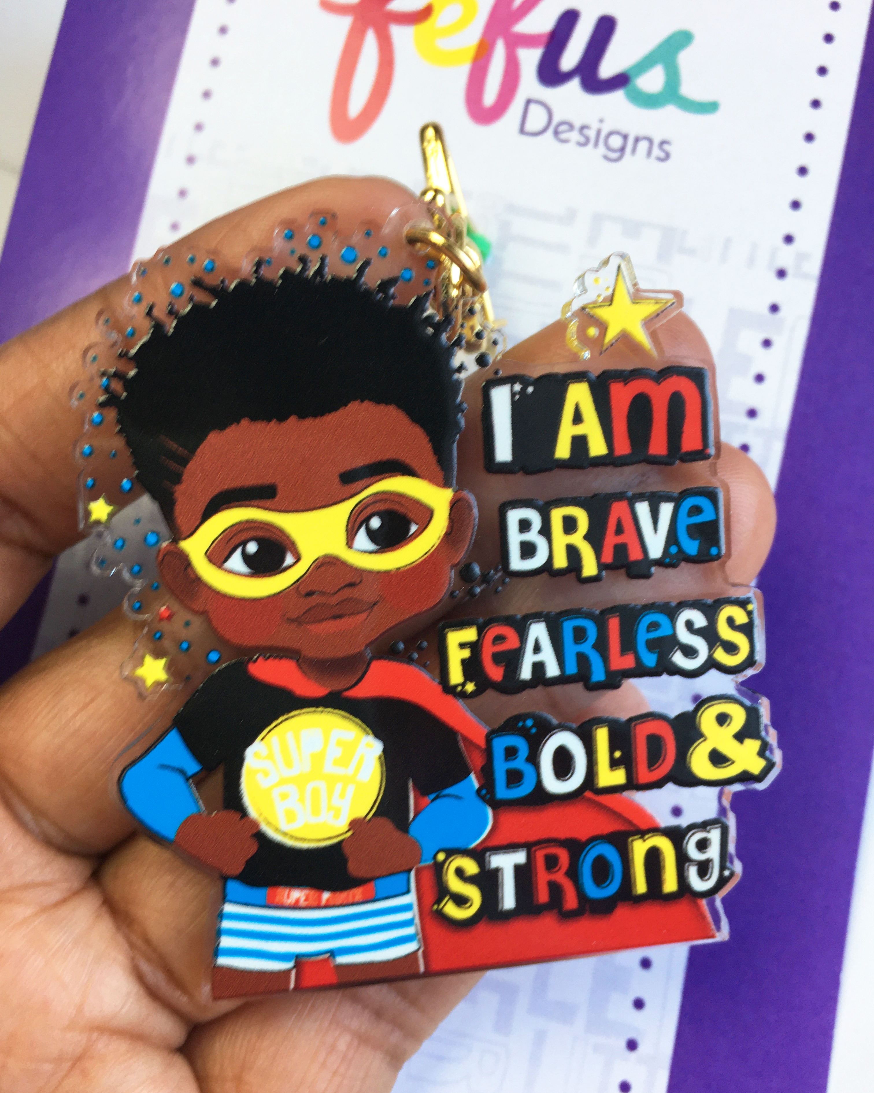 Keyring with a black boy superhero design and text 'I am brave, fearless, bold & strong' held by a hand.