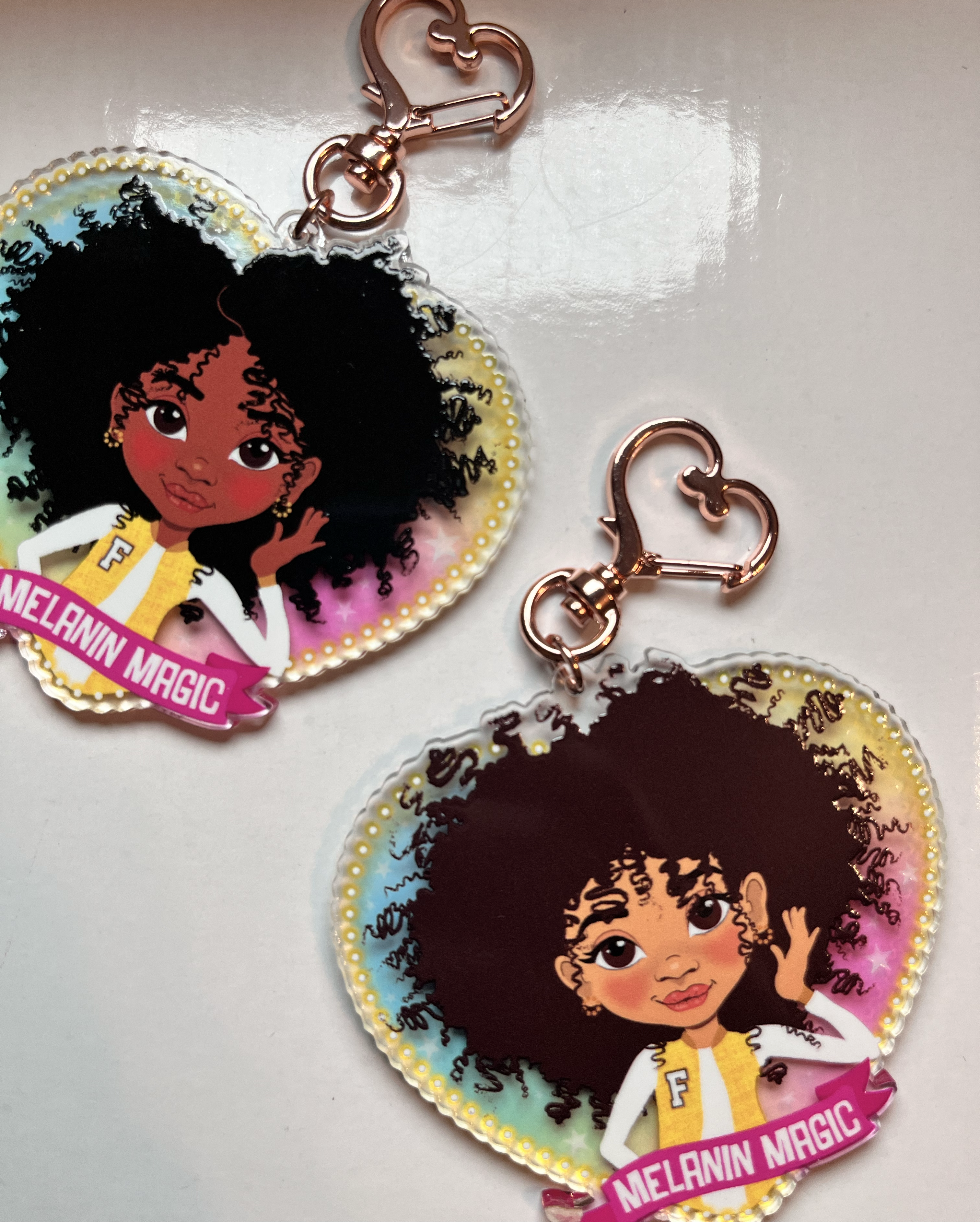 Two keychains with illustrations of a girl with an afro and 'Melanin Magic' text on a transparent rainbow glazed background.