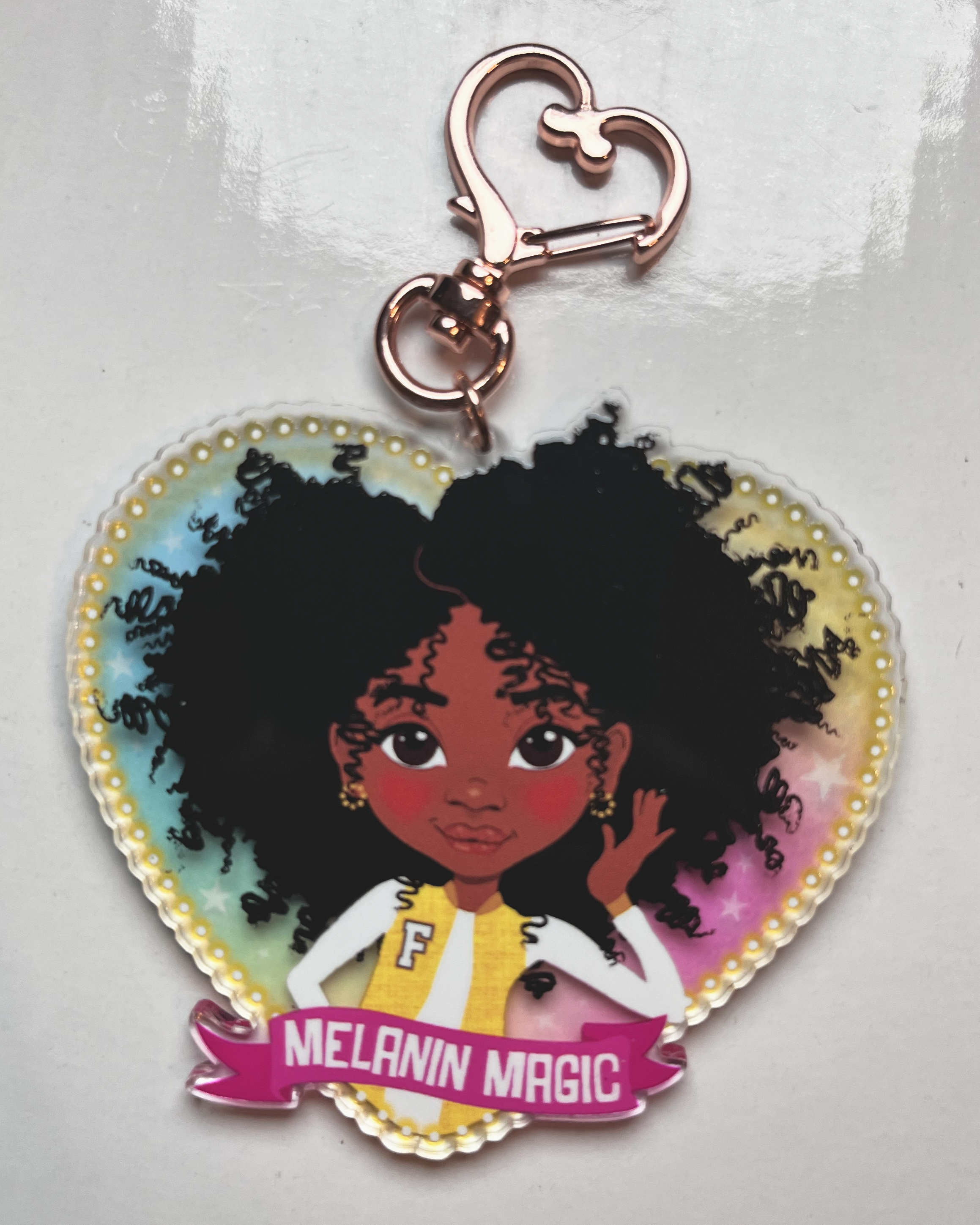 Heart-shaped keychain with an illustration of a black girl large coily afro hair. The text 'Melanin Magic' set on a pink scroll banner. Rainbow glazed background with yellow glow lights around the edge