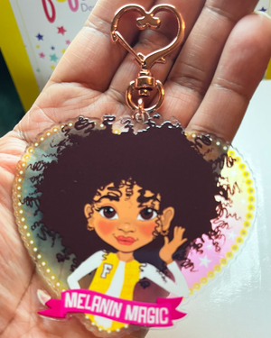 Heart-shaped keychain with a Mixed race Afro Girl hand drawn character and 'Melanin Magic' text on a banner scroll. Brown tween / teen Girl keyring with curly afro and “Melanin Magic” affirmation