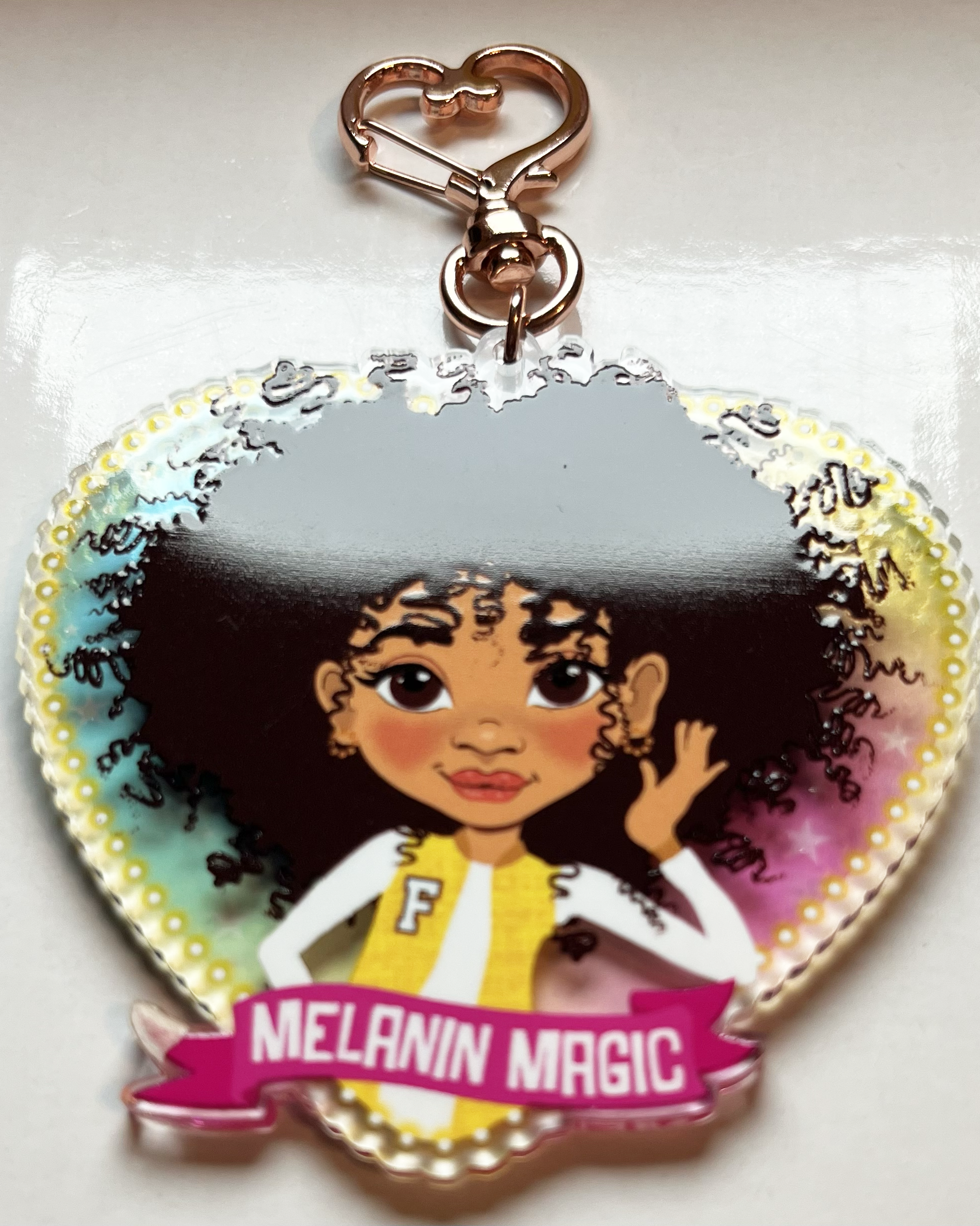 Heart-shaped keychain with a brown afro girl cartoon character and 'Melanin Magic' text on a transparent rainbow background