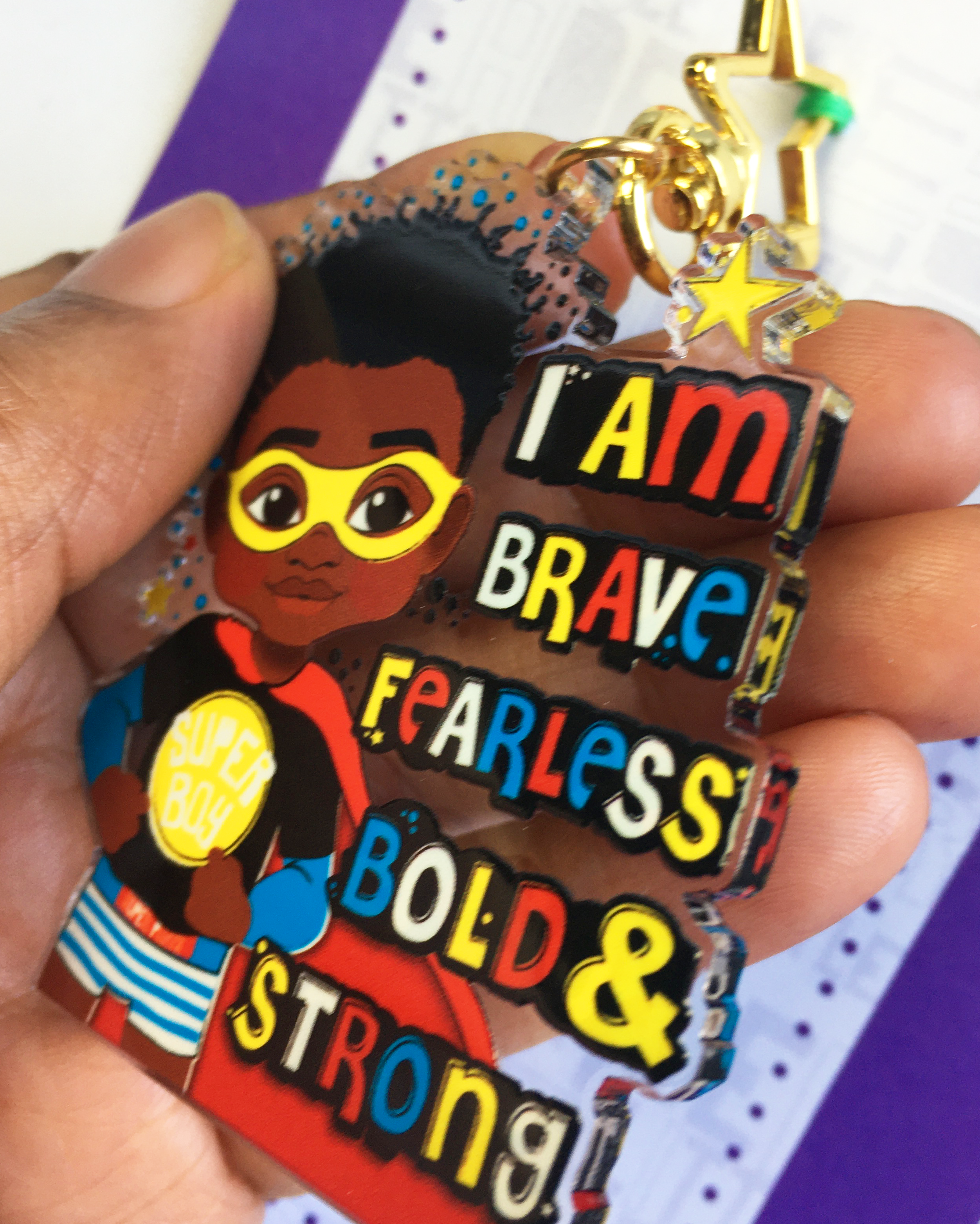 sideview of a black boy superhero keychain with motivational text held by a hand against a colourful background. ouble-sided superhero affirmation bag charm showing vibrant colours and glossy finish, 63.5mm, by Fefus Designs