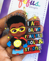Black boy superhero design keyring featuring a confident child with curly hair and a cape by Fefus Designs. Text 'I am brave, fearless, bold & strong' held by a hand.