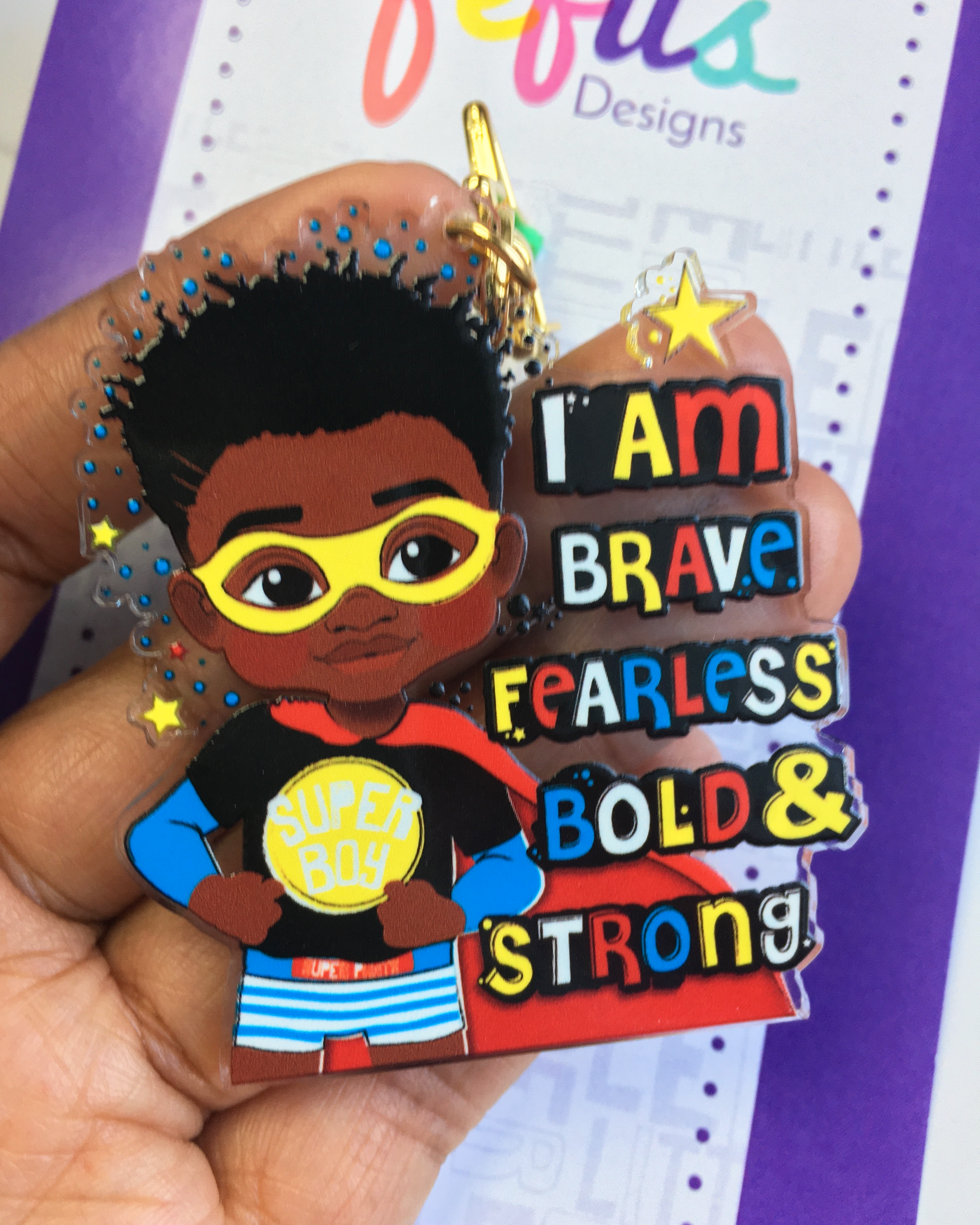 Keyring with a child character wearing sunglasses and a cape, with text 'I am brave fearless bold & strong' held by a hand.