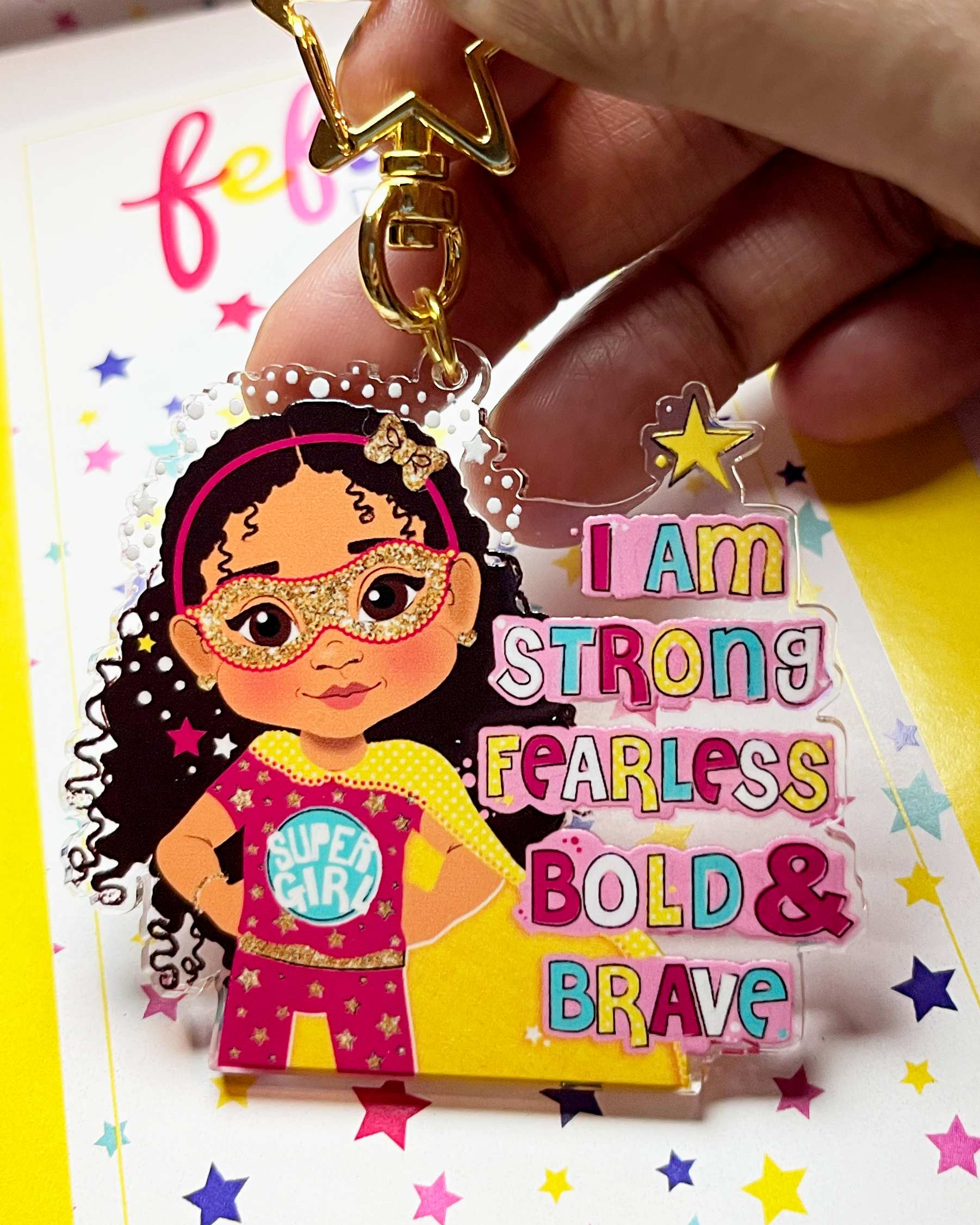 Keychain with a mixed race girl character wearing a superhero outfit and 'I am strong fearless bold & brave' colourful text on a pink background.