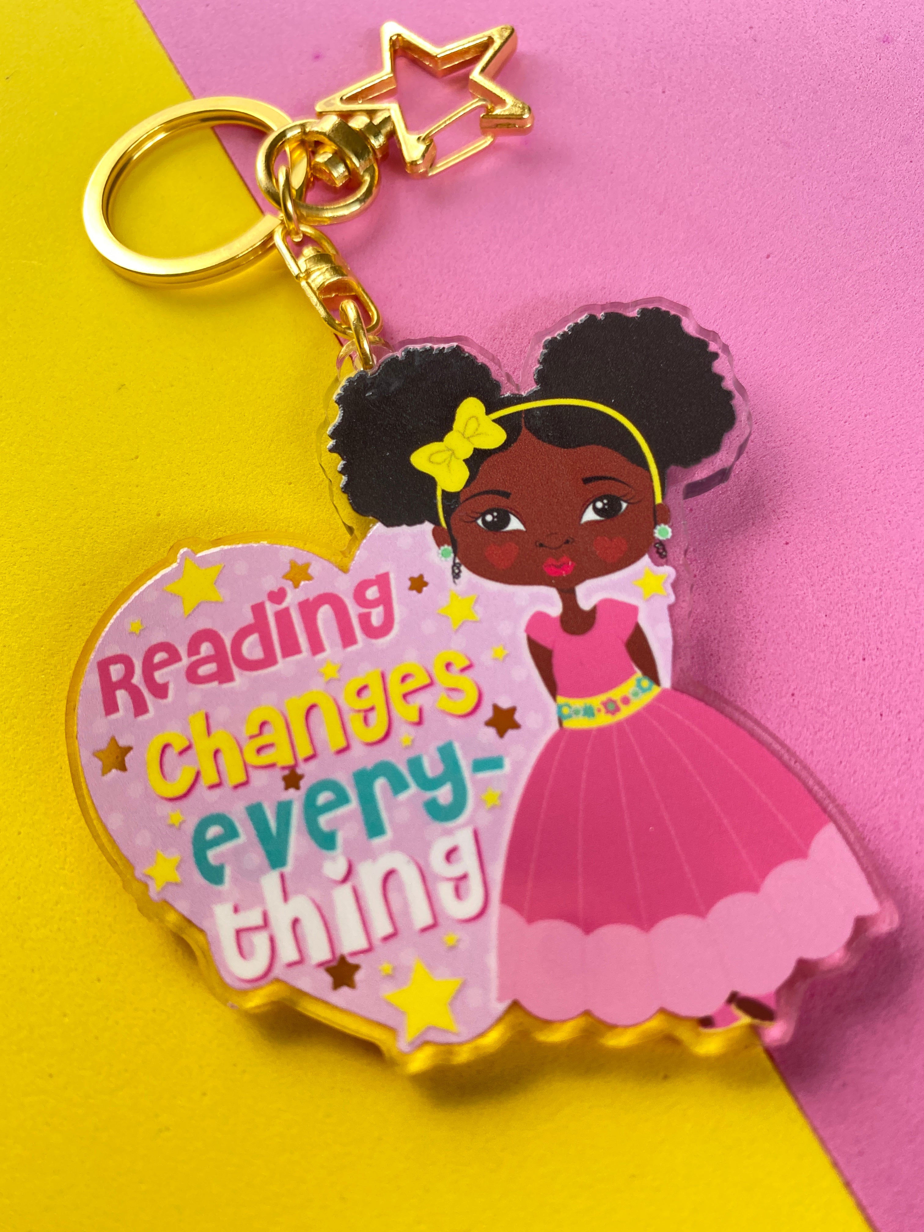 A pink-colored acrylic keyring with a gold-plated star-shaped keyring and a printed design featuring a character and the text 'Reading Changes Everything'.